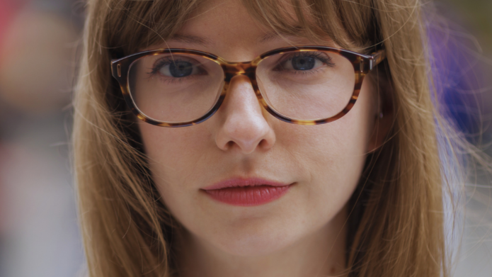 Portret młodej kobiety z długimi, jasnobrązowymi włosami i grzywką, noszącej okulary w brązowej oprawce w stylu retro. Kobieta patrzy prosto w obiektyw z delikatnym, neutralnym wyrazem twarzy. Tło jest rozmyte, co sugeruje, że zdjęcie zostało wykonane na zewnątrz.
