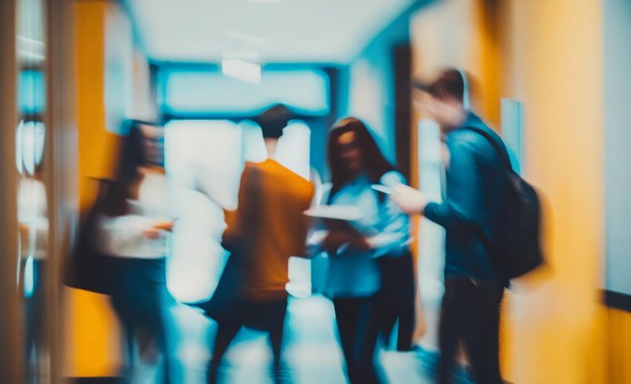 Rozmyte zdjęcie przedstawiające grupę młodych osób stojących w korytarzu uczelni lub biura. Postacie w ruchu rozmawiają lub przeglądają dokumenty. Ciepłe odcienie żółci i błękitu nadają scenie dynamiczny, nowoczesny charakter, oddając atmosferę codziennych spotkań i wymiany pomysłów w środowisku akademickim.