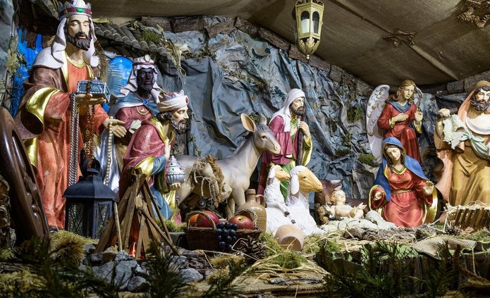 Na obrazie widoczna jest szopka bożonarodzeniowa z wieloma figurami. Scena przedstawia narodzenie Jezusa. W środku leży małe dziecko – Jezus, ułożone na jasnym sianie w drewnianym żłóbku. Dziecko ma jasną skórę, jest nagie, owinięte delikatną tkaniną. Ręce ma lekko uniesione. Po prawej stronie dziecka klęczy Maryja. Ma spokojną twarz i spuszczony wzrok. Jest ubrana w czerwoną suknię i niebieski płaszcz. Ręce ma złożone w geście modlitwy. Obok Maryi stoi święty Józef. Jest ubrany w jasną szatę, ma brodę i trzyma baranka na rękach. Jego twarz jest łagodna. Po lewej stronie znajdują się Trzej Królowie. To trzej mężczyźni w bogatych, kolorowych strojach. Jeden z nich klęczy i trzyma naczynie z darem. Pozostali stoją, trzymając prezenty dla dziecka. Jeden z Królów ma ciemną skórę, pozostali jaśniejszą. Za nimi widać zwierzęta: osła, owce i baranki. Zwierzęta stoją spokojnie, zwrócone w stronę dziecka. W tle znajduje się skalna grota i fragmenty drewnianej konstrukcji. Nad postaciami wiszą lampy i gwiazdy, które oświetlają scenę ciepłym światłem. Całość ma uroczysty i spokojny charakter, a kolory są ciepłe: brązy, złoto, czerwień i niebieski. Scena symbolizuje narodziny Jezusa i moment oddania czci przez ludzi i zwierzęta.