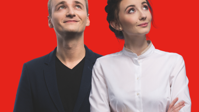 "Młody mężczyzna i kobieta stoją obok siebie na tle intensywnie czerwonego tła, oboje patrzą w górę z lekkimi uśmiechami. Mężczyzna ubrany jest w ciemną marynarkę i czarną koszulkę, a kobieta ma na sobie elegancką, białą koszulę z kołnierzykiem i skrzyżowane ręce."