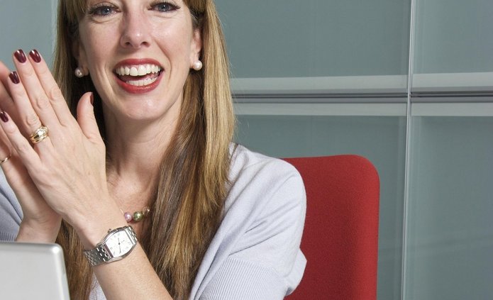 kobieta siedząca na bordowym krześle przy komputerze. Blondynka, długie proste włosy, ubrana w biała koszulkę, bije brawo i jest uśmiechnięta