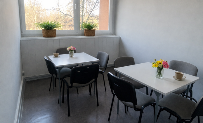 Na zdjęciu widoczny jest jasny, niewielki pokój socjalny. W pomieszczeniu stoją dwa białe stoliki, a przy każdym z nich ustawione są cztery krzesła w kolorze szarym lub czarnym. Na stolikach znajdują się małe wazoniki z kolorowymi kwiatami oraz filiżanki. Na końcu pomieszczenia znajduje się duże okno, przez które wpada dużo naturalnego światła. Na parapecie stoją dwie doniczki z zielonymi roślinami. Za oknem widać drzewa bez liści, co może wskazywać na porę jesienną lub wczesną wiosnę. Ściany pokoju są białe, a podłoga w odcieniu szarości. Na suficie zamontowana jest długa lampa sufitowa. Pomieszczenie wygląda na uporządkowane, spokojne i przygotowane do odpoczynku lub rozmowy przy stoliku.