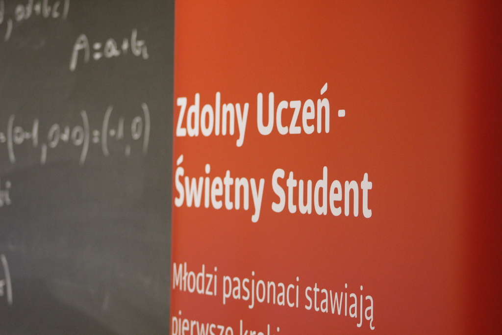 Na zdjęciu jest czerwony baner z białymi napisami: Zdolny uczeń - świetny student. W tle jest czarna tablica z zapisanym działaniem matematycznym