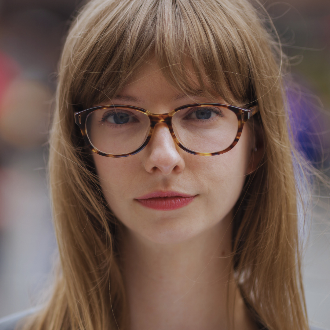 Portret młodej kobiety z długimi, jasnobrązowymi włosami i grzywką, noszącej okulary w brązowej oprawce w stylu retro. Kobieta patrzy prosto w obiektyw z delikatnym, neutralnym wyrazem twarzy. Tło jest rozmyte, co sugeruje, że zdjęcie zostało wykonane na zewnątrz.