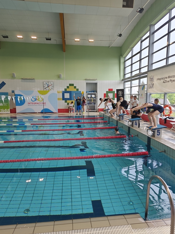 Wnętrze krytej pływalni podczas zawodów pływackich. Kilku zawodników w strojach kąpielowych stoi na słupkach startowych, przygotowując się do skoku do wody. Jeden z zawodników jest już w trakcie skoku. 