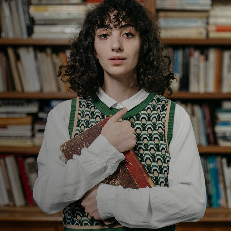 Młoda kobieta o ciemnych, kręconych włosach stoi na tle regału z książkami. Ubrana jest w białą koszulę i zielony sweter w geometryczny wzór. Trzyma w ramionach starą, oprawioną książkę i patrzy spokojnie w obiektyw.